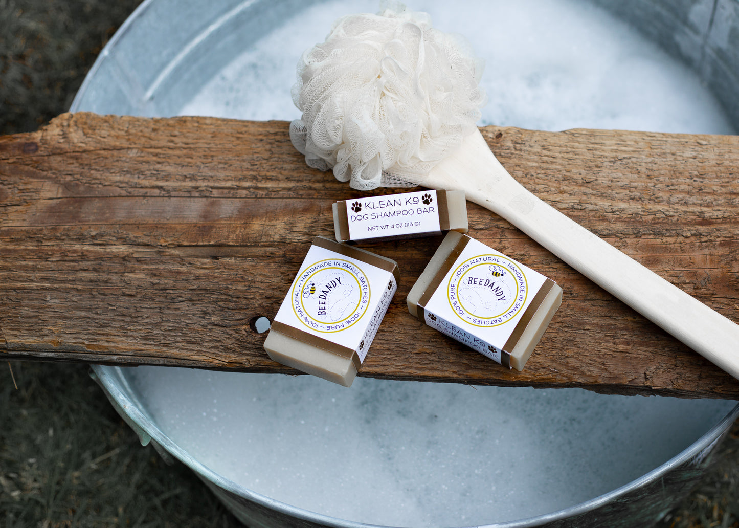 Beedandy Klean K9 dog shampoo bars on a wooden plank above a metal tub filled with water and a bath brush.