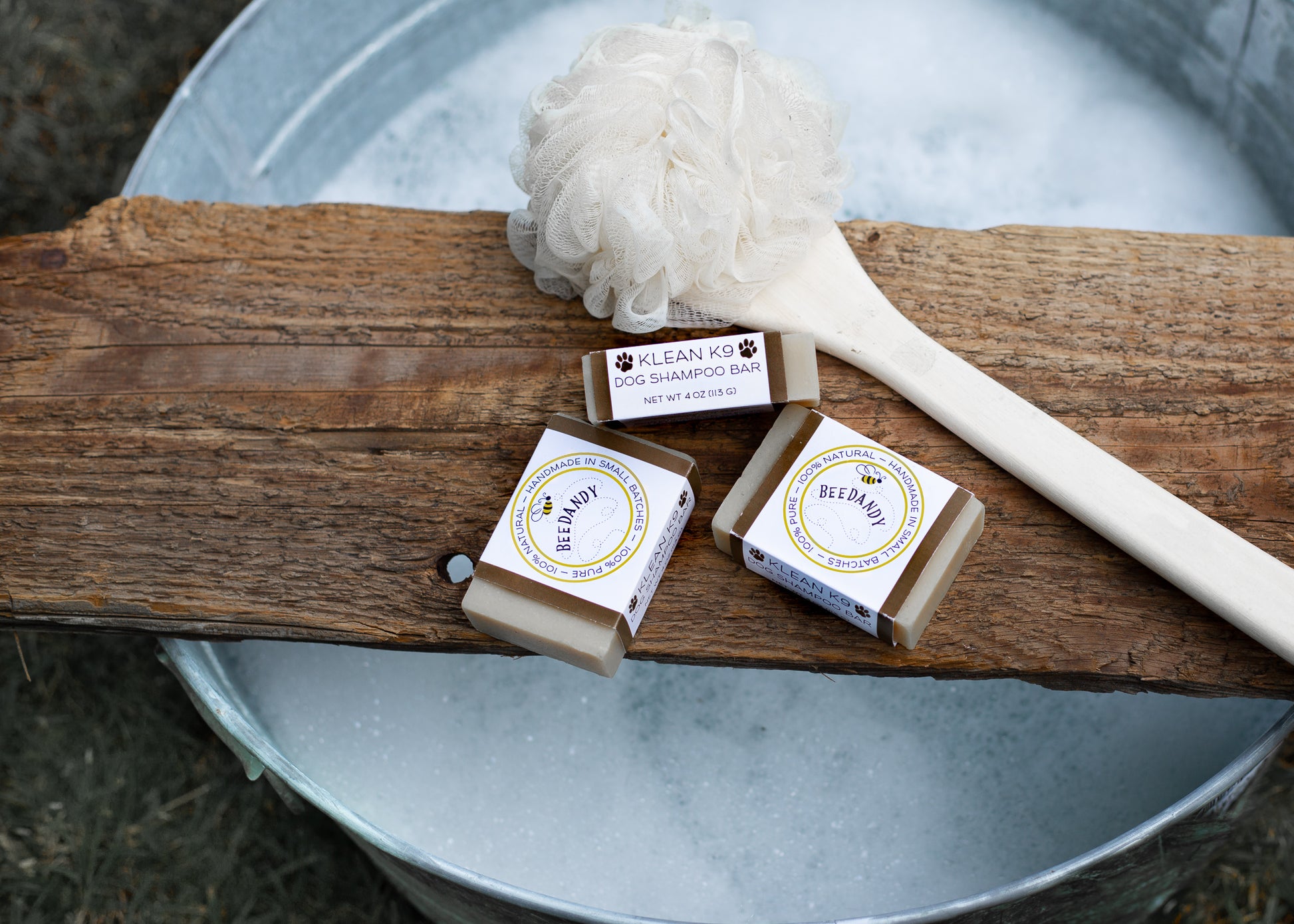 Beedandy Klean K9 dog shampoo bars on a wooden plank above a metal tub filled with water and a bath brush.