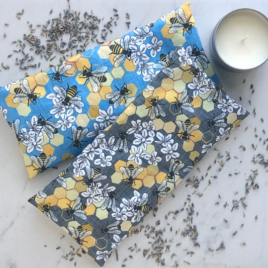 Two patterned fabric pillows with bee and honeycomb designs on a light surface, surrounded by dried lavender, next to a candle.