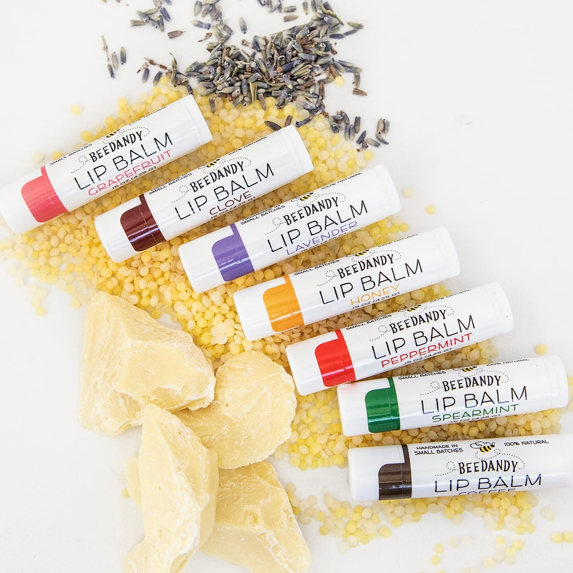 Seven Beedandy lip balm tubes in different flavors displayed on a bed of yellow beeswax pellets with cocoa butter chucks and lavender buds