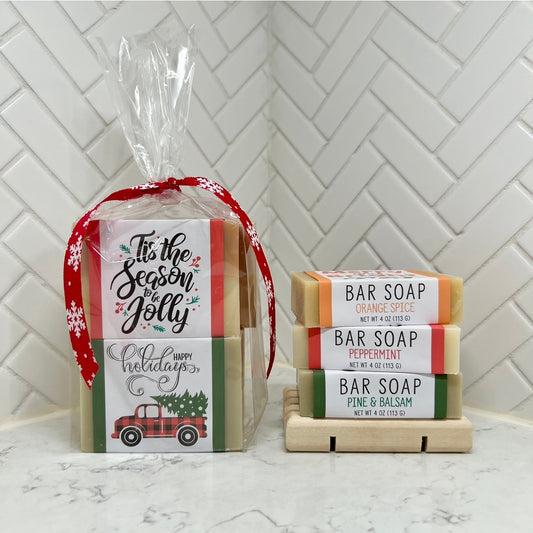 Holiday gift set of wrapped bar soaps tied with a red ribbon, displayed next to three stacked bar soaps labeled Orange Spice, Peppermint, and Pine & Balsam on a wooden soap dish.