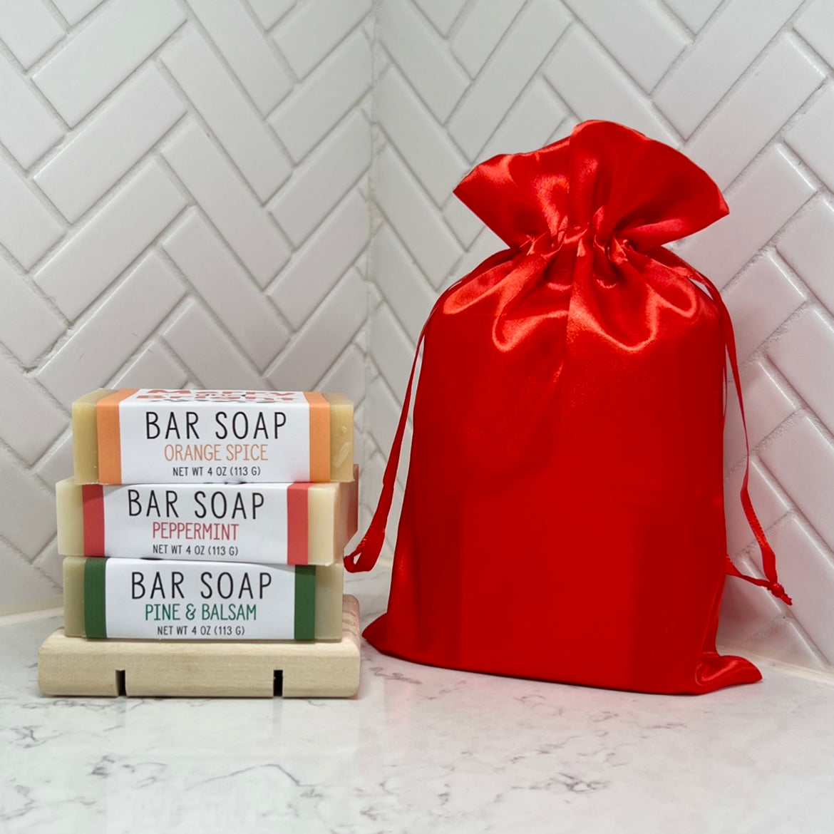 Three stacked bar soaps labeled Orange Spice, Peppermint, and Pine & Balsam on a wooden soap dish, next to a red satin bag.