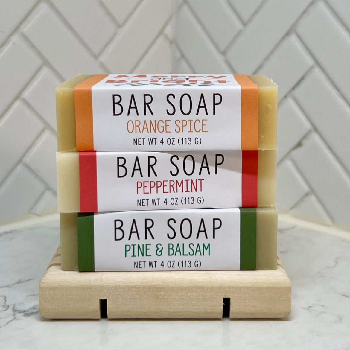 Three stacked bar soaps labeled Orange Spice, Peppermint, and Pine & Balsam on a wooden soap dish.