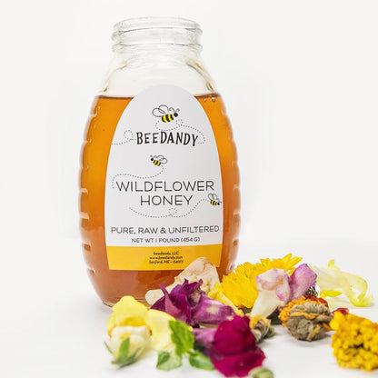 A glass jar of raw, unfiltered honey labeled 'WILDFLOWER HONEY' from BeeDandy, surrounded by an assortment of local wildflowers.