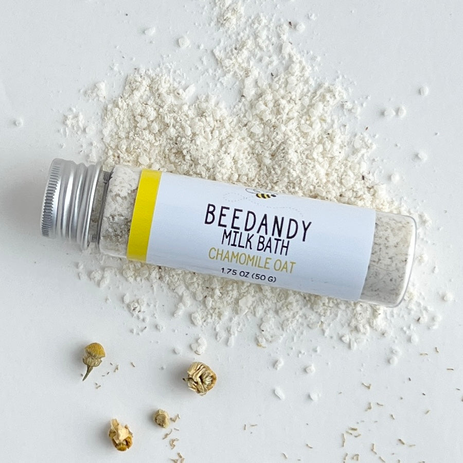 A clear tube of milk bath labeled chamomile oat with white label and silver cap. Against a light grey background and light yellow powder.