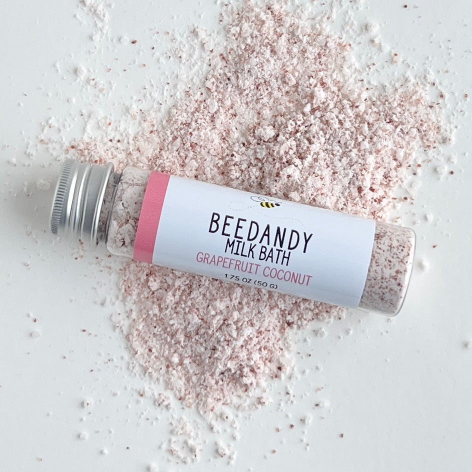 A clear tube of milk bath labeled grapefruit coconut with white label and silver cap. Against a light grey background and some pink powder. 